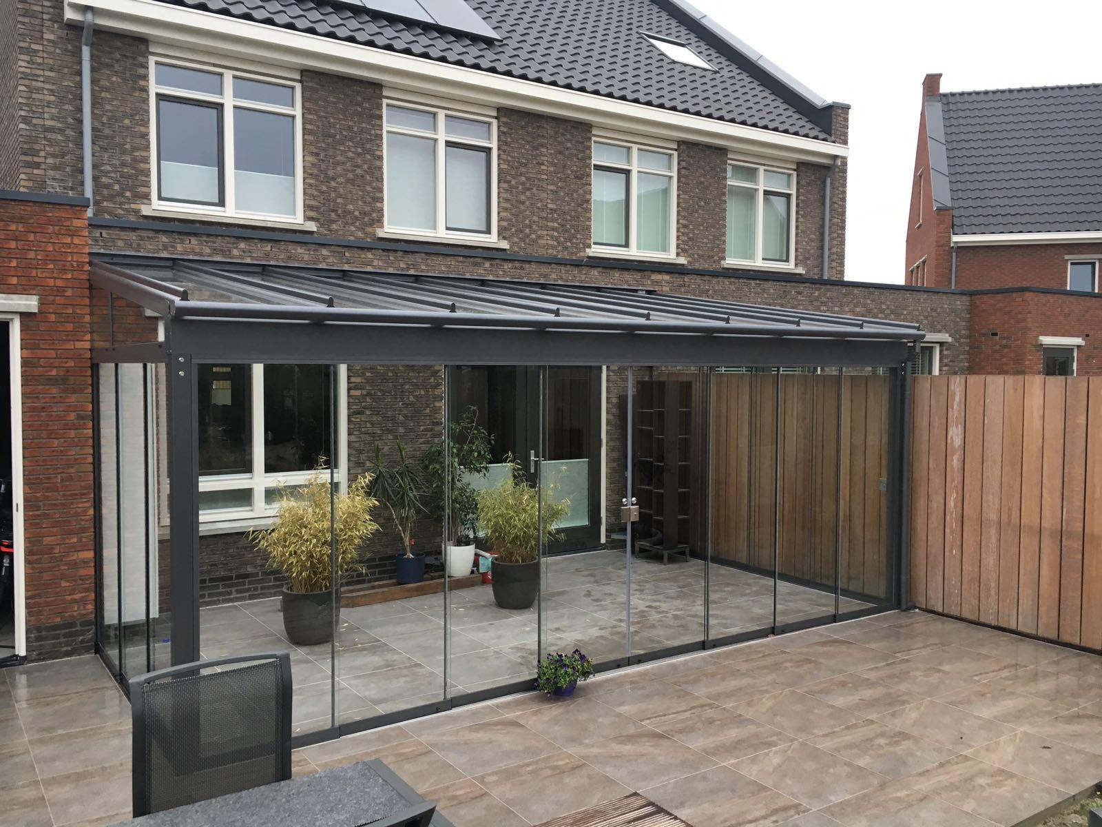 Een moderne woning met een terrasoverkapping op maat met een glazen omheining, potplanten, tegelvloer en houten schutting. De buitenkant heeft grote ramen en een donker dak.