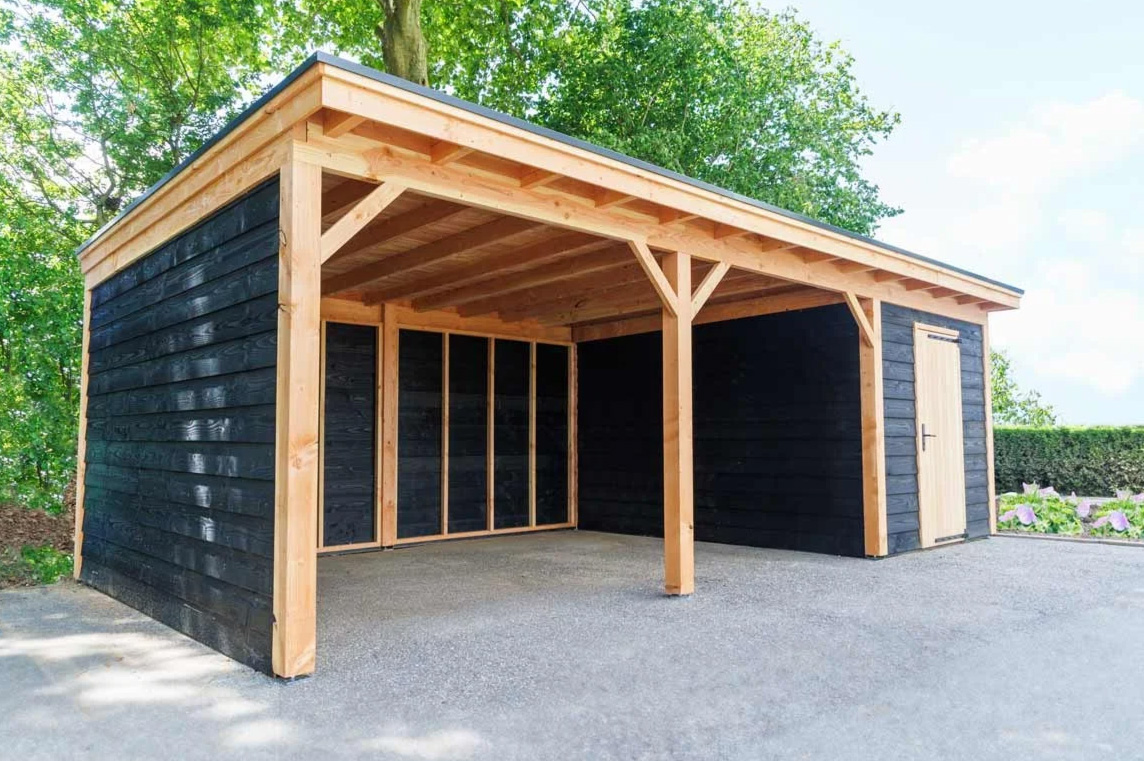 Een houten carport met een plat dak, aan twee kanten open, met zwarte horizontale planken en een frame van natuurlijk hout, met een aangrenzende berging en een lichtgekleurde deur, buiten gelegen in de buurt van bomen.