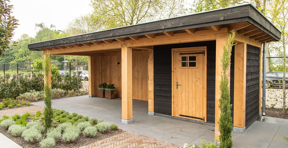 Een modern houten tuinhuisje met een houten overkapping naast een gesloten deur, omgeven door netjes aangelegde beplanting en struiken, met bomen op de achtergrond.