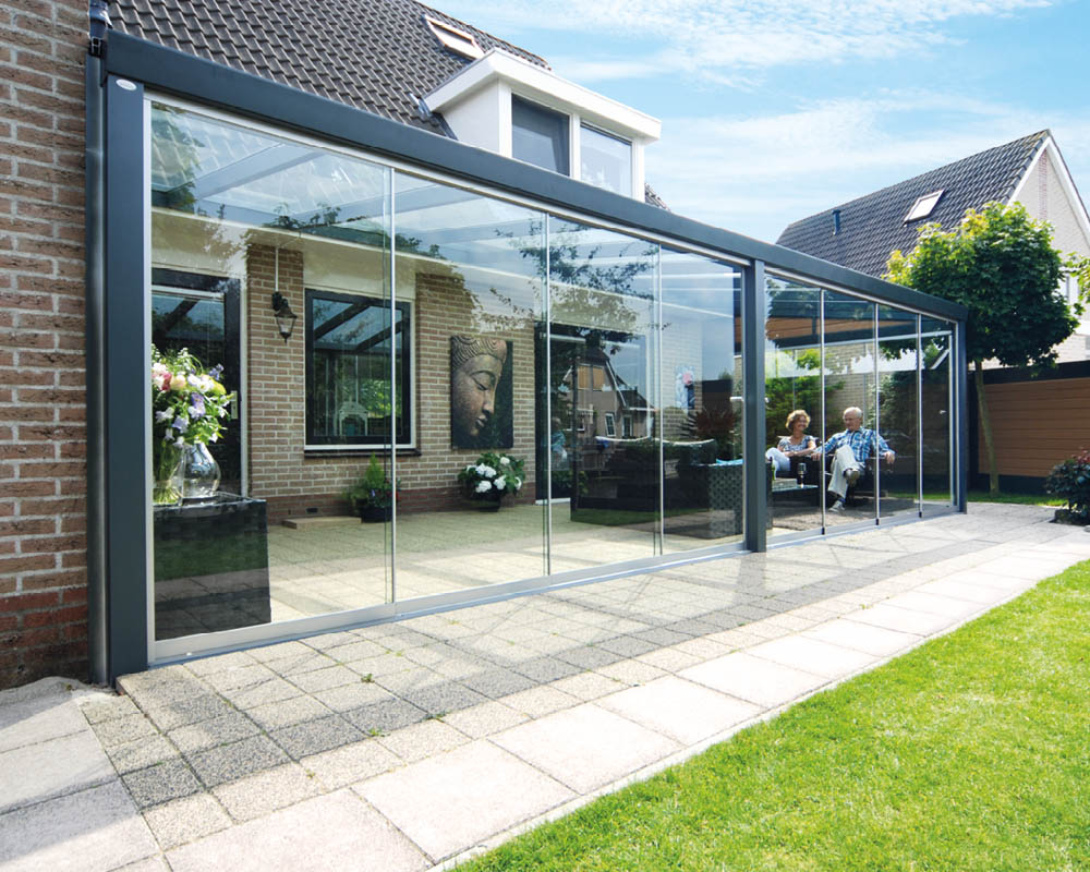 Een moderne glazen terrasoverdekking aan een huis, met twee mensen die binnen aan een tafel zitten. De overdekking heeft een tegelvloer en is omgeven door een groen gazon en een betegeld voetpad.