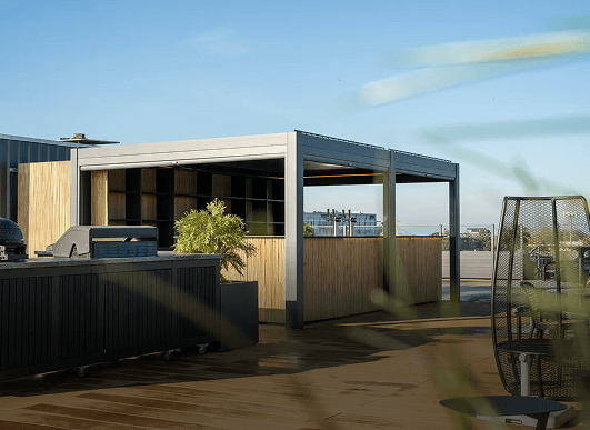 Modern dakterras met een schaduwrijke pergola, buitenkeuken en zitgedeelte. De ruimte is beplant en kijkt uit over een stadsgezicht onder een strakblauwe hemel.