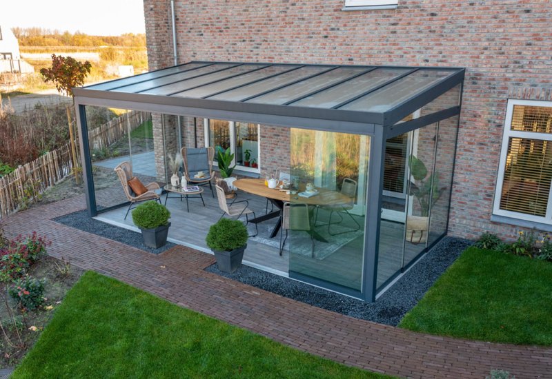 Een moderne patio met glazen omheining aan een bakstenen huis, met buitenmeubilair, planten in potten en een houten vloer, omgeven door een groen gazon en een stenen looppad.