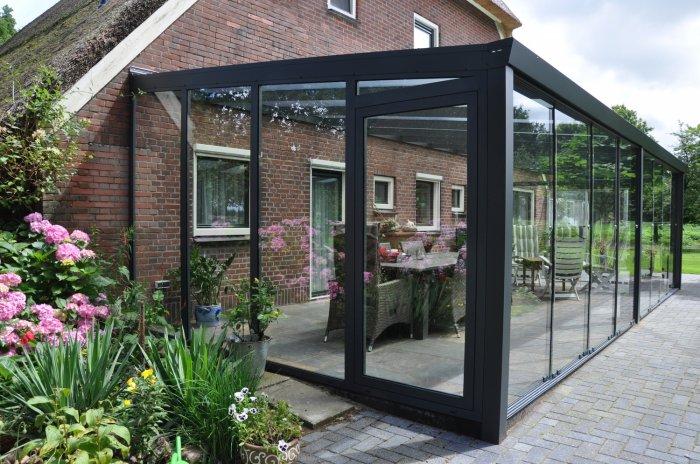 Een moderne glazen serre met een zwart frame is vastgemaakt aan een bakstenen huis, met uitzicht op een tuin met roze bloemen en groen. Binnen staan een tafel en stoelen klaar om aan te dineren of te ontspannen.