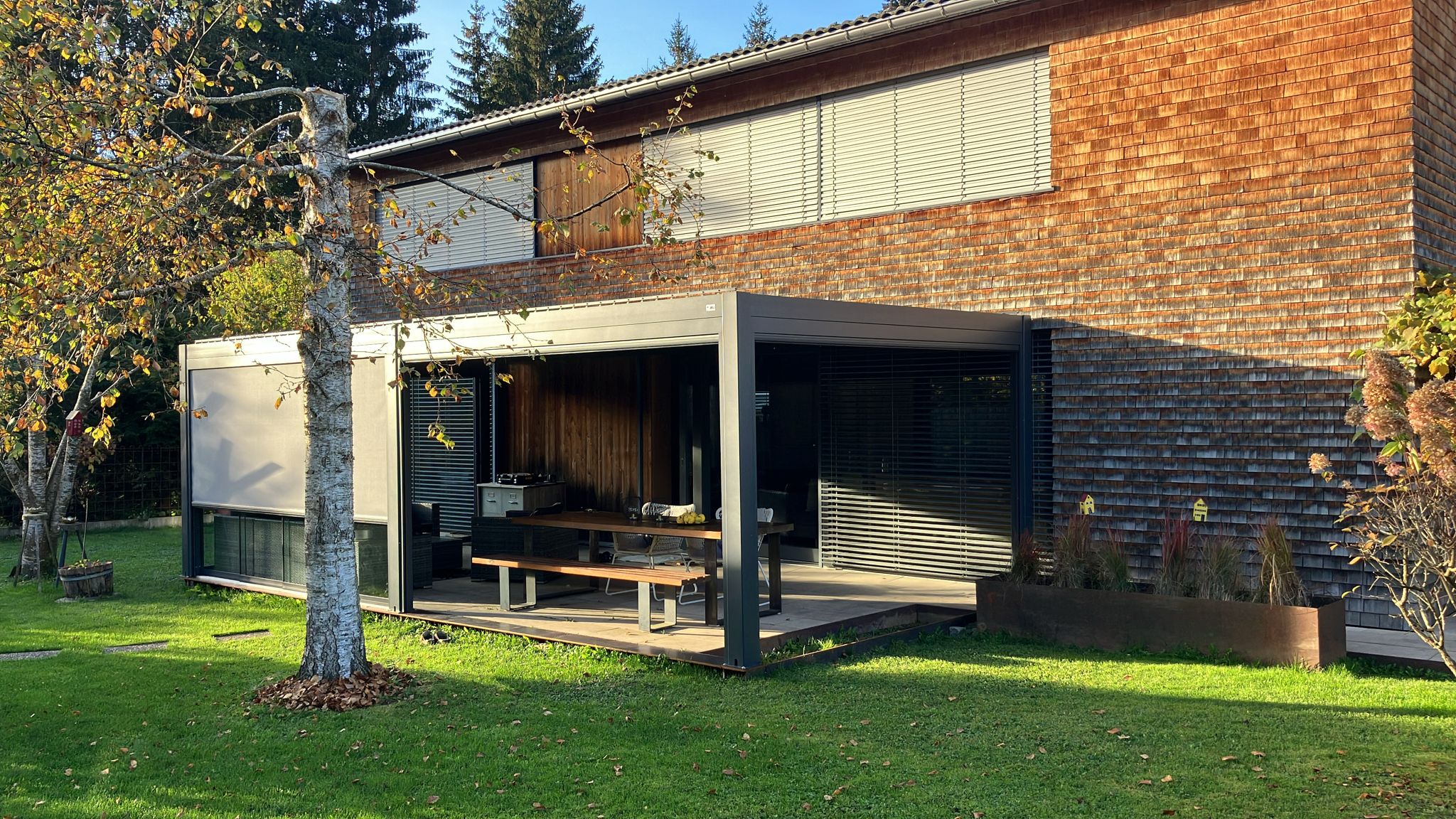 Een modern huis met houten gevelbekleding heeft een overdekt terras (overkapping), een eettafel buiten, een grill en een groen gazon met een boom op de voorgrond. Het strakke zwarte frame biedt stijlvolle schaduw om te ontspannen of te entertainen.