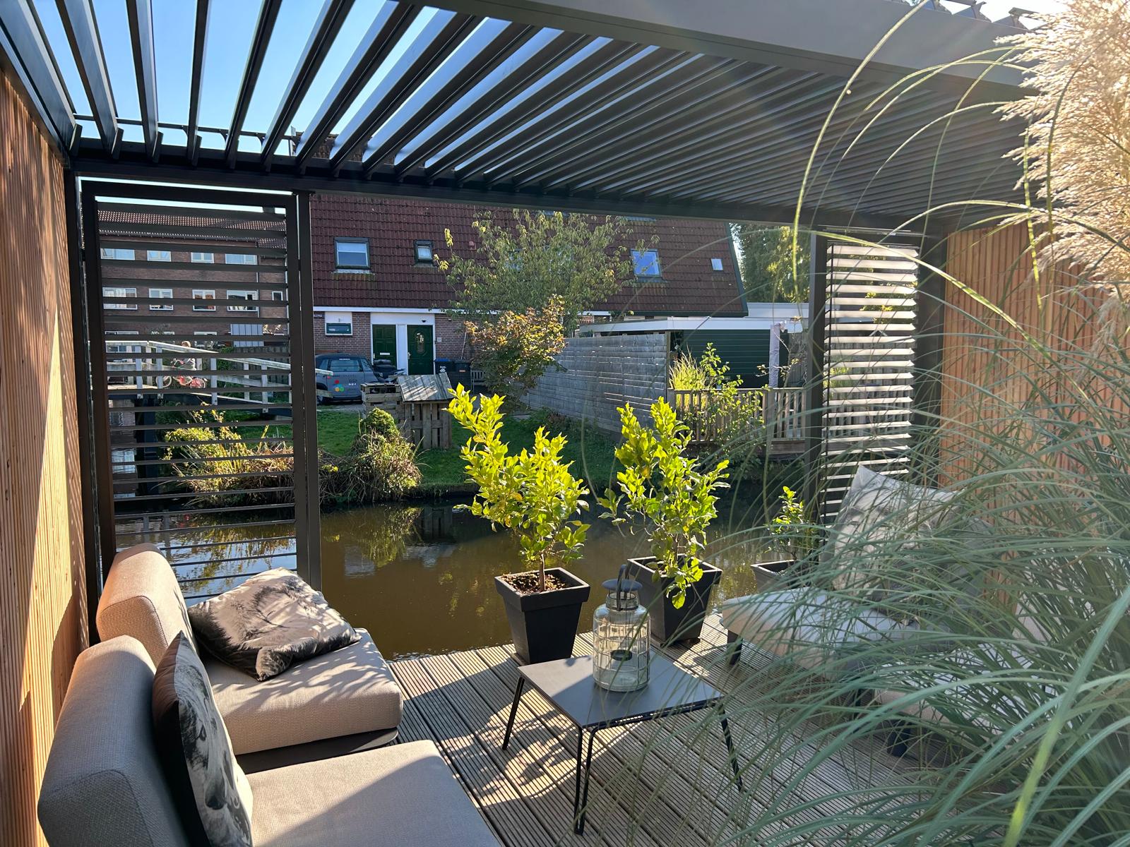 Een gezellig buitenterras met moderne stoelen, planten in potten en een overkapping met latten kijkt uit over een klein kanaal en woonhuizen op de achtergrond, waar het zonlicht naar binnen stroomt.