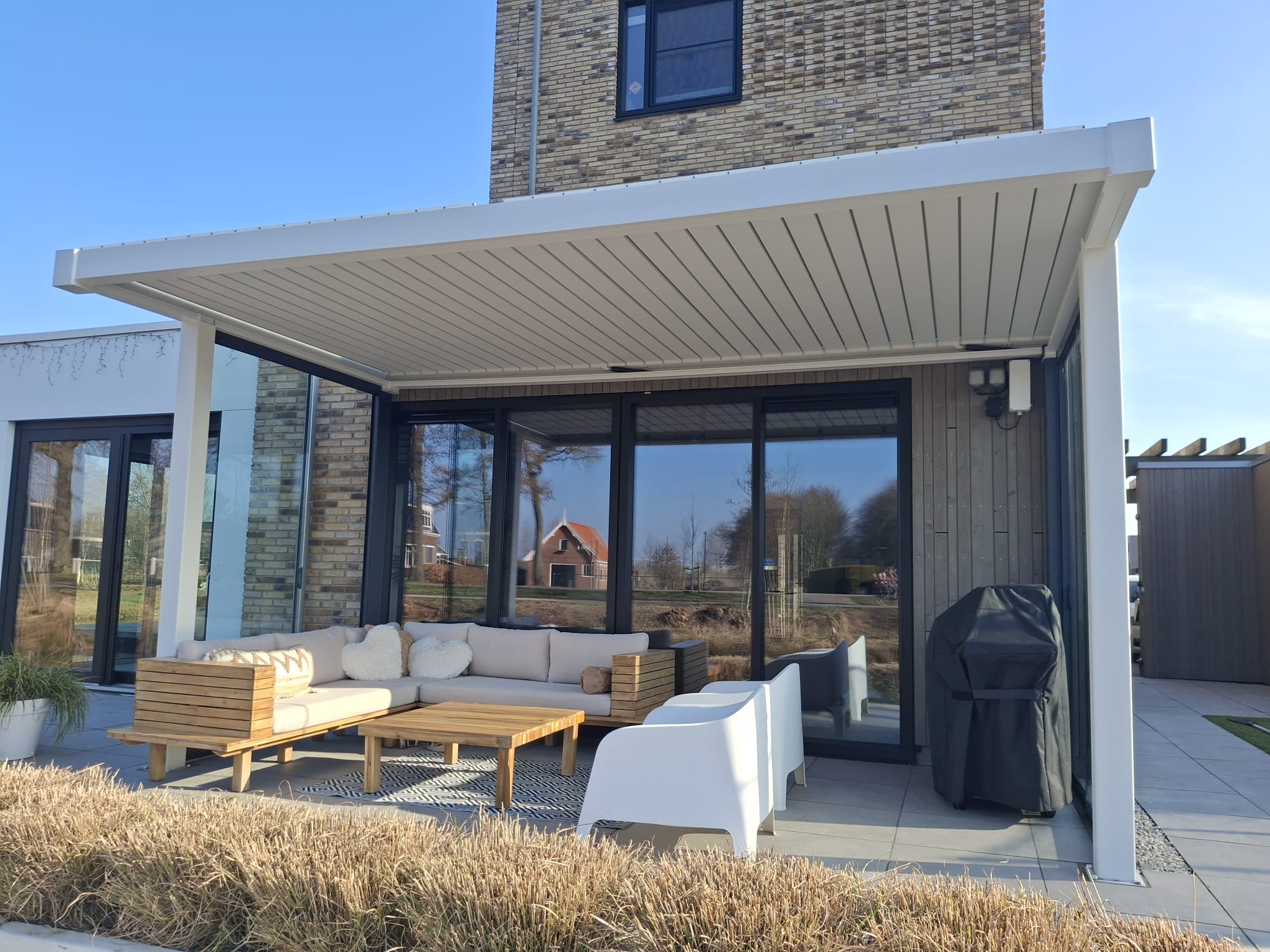 Een moderne patio met een witte overkapping, buitenbank, fauteuils en een grill, tegen grote glazen deuren en ramen van een hedendaags huis, met droog siergras op de voorgrond.