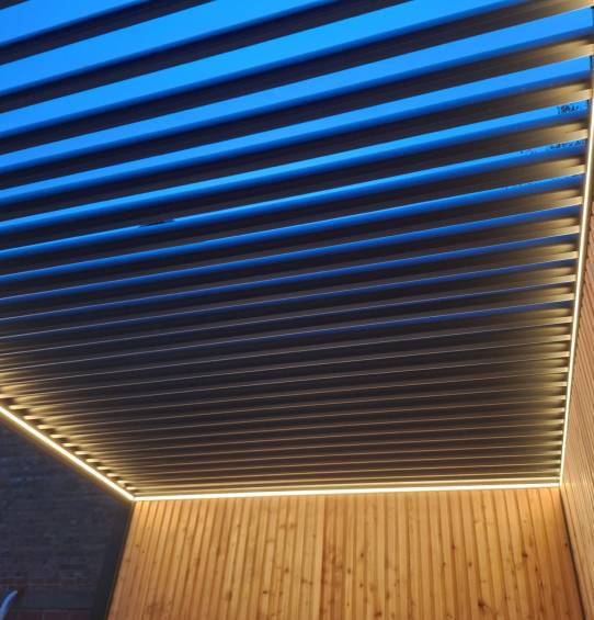 Een modern plafond met parallelle metalen lamellen en geïntegreerde LED-stripverlichting langs de randen, boven wanden met houten panelen, tegen een helderblauwe lucht.