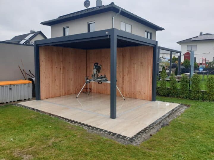 Een modern tuinhuisje met open zijkanten, een tegelvloer en houten wanden, staat op een gazon en lijkt op een lamellenoverkapping. In het midden staat een elektrische zaag op een standaard, met woonhuizen zichtbaar op de achtergrond.