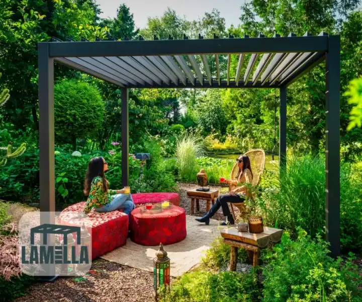 Twee mensen zitten onder een moderne pergola in een weelderige tuin, omringd door rode, gepatroneerde ronde zitplaatsen en houten tafels. Een lantaarn rust op de grond. Zonlicht filtert door bomen en werpt schaduwen. Een Lamella-logo staat in de linkerbenedenhoek.