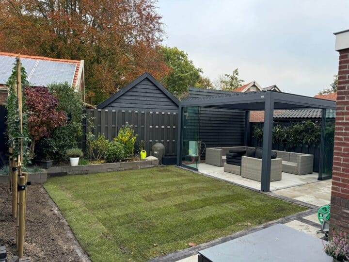 Een achtertuin met pergola verse graszoden en een lamellen overkapping die het moderne terras aan de rechterkant bedekt en veelzijdige schaduw biedt. Een donkergrijze schuur staat op de achtergrond, terwijl potplanten en bomen weelderig groen toevoegen. De lucht is bewolkt.