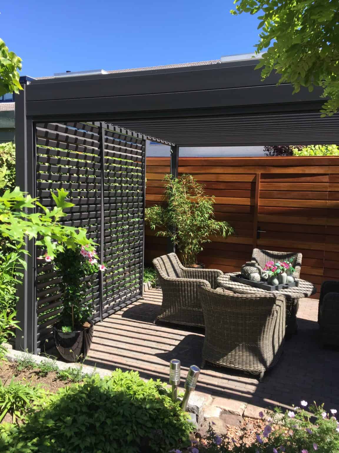 Buitenpatio met rieten stoelen en een tafel onder een pergola. Zonlicht werpt schaduwen door het lamellendak. Omringd door groen en een houten hek, biedt de ruimte een rustige setting tegen een helderblauwe lucht.