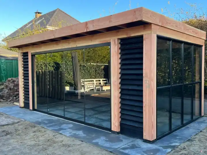Een moderne tuinkamer met grote glazen panelen en houten accenten. De structuur heeft zwarte lattenbekleding, omgeven door een geplaveid terras. Een houten picknicktafel is zichtbaar op de achtergrond.