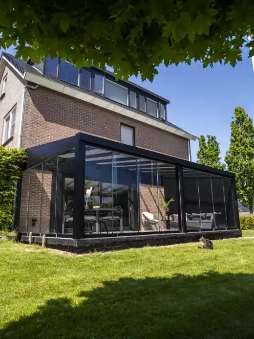 Een modern huis heeft een glazen patio met Terrasoverkapping glas, zwart metalen frame, een eethoek en een bank. Het gebied beschikt over een goed onderhouden gazon, bomen en struiken onder een heldere, zonnige hemel.