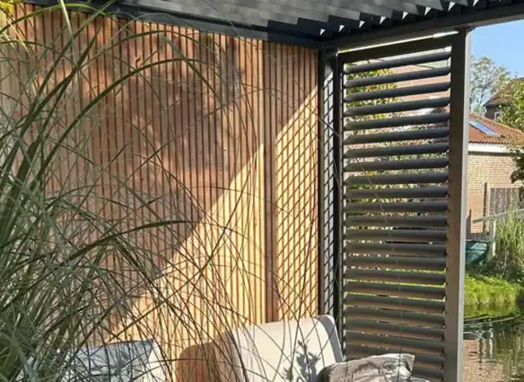 Zitgedeelte buiten met grijze stoelen en een houten lattenwand, gedeeltelijk overschaduwd door metalen lamellen die gestreepte schaduwen werpen. Weelderige groene planten omringen de ruimte en op de achtergrond is een glimp van een huis en tuin te zien.