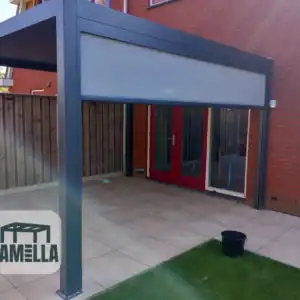 Moderne patio-overkapping bevestigd aan een rood bakstenen huis met deuren met rode omlijsting. De structuur is van metaal met een wit uitschuifbaar scherm. Logo met tekst Lamella verschijnt in de linkerbenedenhoek. Een zwarte emmer staat op de betegelde patiovloer.